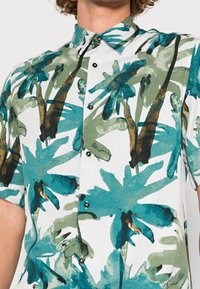 Hombre que lleva una camisa de manga corta, abotonada, con un estampado de palmeras verdes y azules sobre un fondo blanco.