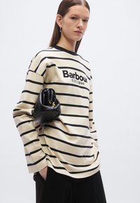 Gestreept beige en zwart shirt met lange mouwen, voorzien van een logo, gecombineerd met een kleine zwarte getextureerde handtas, nonchalant gedragen met een zwarte broek.