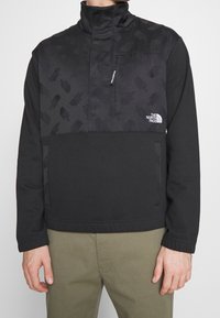 Svart fleecepolo med hög krage, med ett mönstrat övre avsnitt, sidofickor och The North Face-logotyp. Mjuk textur.