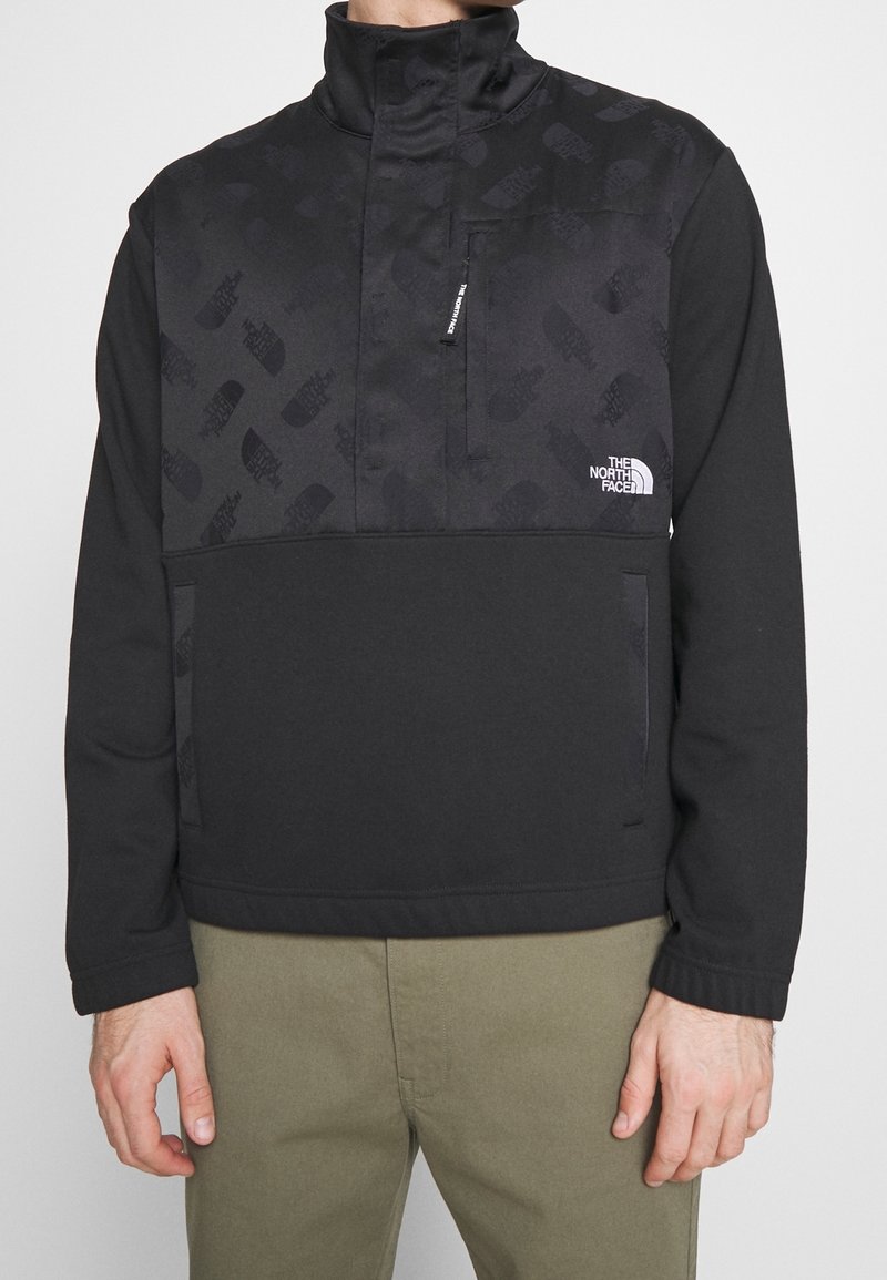 Svart fleecepolo med hög krage, med ett mönstrat övre avsnitt, sidofickor och The North Face-logotyp. Mjuk textur.