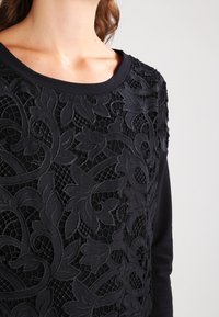 Schwarzes Oberteil mit einem strukturierten Spitzenmuster und floralem Design auf der Vorderseite, vor einem einfarbigen Stoffhintergrund und mit rundem Ausschnitt.