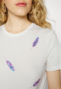 Hvid bomulds t-shirt med farverige palietbesatte fjerdesigns i lilla og blå. Rund halsudskæring og korte ærmer.