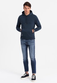 Felpa con cappuccio blu navy con tasca frontale, realizzata in tessuto morbido. Abbinata a jeans azzurri e sneakers scure con lacci crema.
