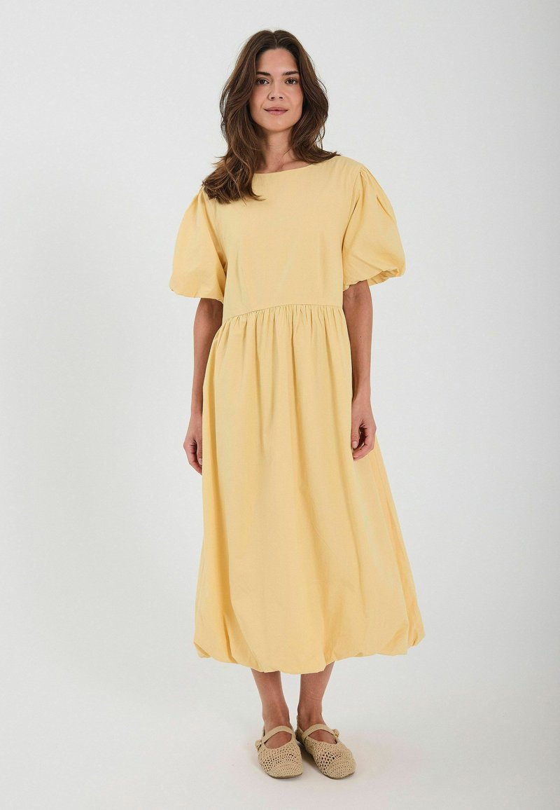 Robe midi en coton jaune avec des manches courtes bouffantes et une taille froncée. Texture lisse et coupe ample, portée avec des chaussures à enfiler tissées beiges.