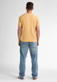 Tricou galben din bumbac cu margini albe la mâneci și la gât, purtat împreună cu blugi lejeri de culoare albastru deschis, cu buzunare la spate și cusături vizibile.