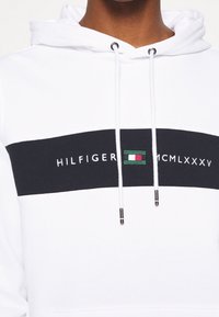 Witte hoodie met zwarte horizontale streep met het "HILFIGER"-logo en groene, witte en rode accenten. Trekkers met zilverkleurige uiteinden.