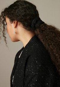 Svart scrunchie med paljetter som håller upp lockigt hår, tillsammans med en texturerad svart jacka. Silverörhängen med en subtil drop-design.