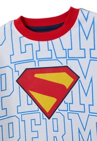 Camiseta de algodón blanca con cuello rojo, que presenta un gran logo de Superman en rojo y amarillo y un texto azul que repite la palabra "PERM".