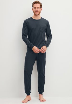 Uomo in piedi a piedi nudi con maglia scura a maniche lunghe con tasca sul petto e pantaloni da jogging coordinati su uno sfondo chiaro uniforme.