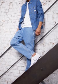 Ljusblå denimjacka med framfickor, parat med ljusblå jeans och vita sneakers, stående på en trappa mot en tegelvägg.