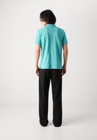 Turquoid póló ing rövid ujjakkal és oldalsó résekkel, fekete nadrággal párosítva, hátulról mutatva. Egyszerű design, látható minták nélkül.