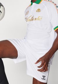 Bílý fotbalový dres s krátkým rukávem, zelenými a oranžovými akcenty, se zlatým nápisem "Authentic" a číslem "10" na přední straně. Přítomné logo Kappa.