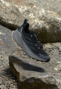 Chaussure de sport noire et grise avec une tige en mesh, une semelle en caoutchouc texturée et des lacets réfléchissants, comportant trois bandes sur le côté et une languette au talon.