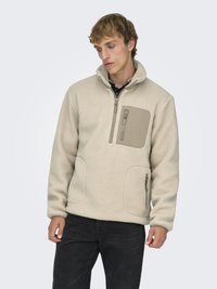 Mladić u bež flis jakni s patentnim zatvaračem na prsima i bočnim džepovima, uparenom s crnim hlačama, stoji na svijetloj pozadini.