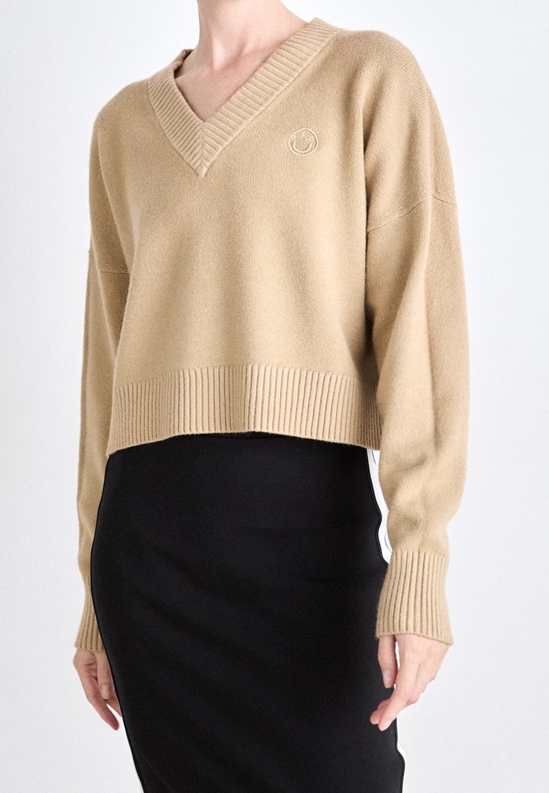 Pull court beige à col en V avec manches longues côtelées, porté sur une jupe noire avec des bandes blanches sur les côtés.