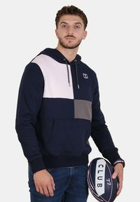 Homme portant un sweat à capuche coloré bleu marine, rose et gris tenant un ballon de rugby avec le texte "CLUB 77".
