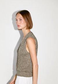 Junge Frau mit kurzen braunen Haaren, die eine ärmellose braune Strickweste und einen weißen Rock trägt, mit dem Gesicht nach links vor einem schlichten weißen Hintergrund.