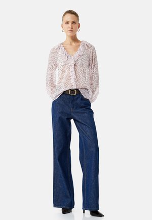 Mujer con blusa rosa transparente de lunares con volantes, vaqueros azul de talle alto y pata ancha, cinturón negro y tacones negros puntiagudos.