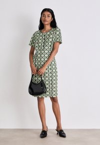 Kurzes Kleid mit grünem und schwarzem geometrischem Muster, tailliert geschnitten, ergänzt durch eine schwarze glänzende Handtasche und schlichte hohe Clogs.