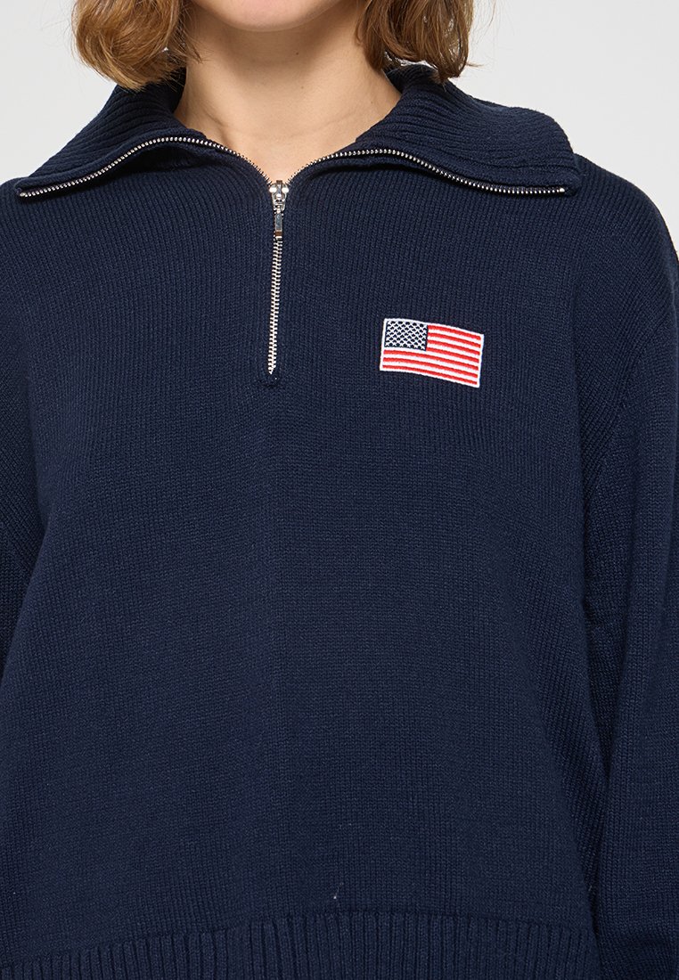 Suéter azul marino con cuello de media cremallera, textura acanalada y una bandera americana bordada en el pecho. Diseño de tejido suave y de punto.