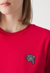 Mujer vistiendo una camiseta roja con un pequeño parche brillante negro y plateado de un rostro estilizado con gafas de sol en el pecho.