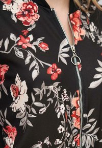 Veste bomber noire à fleurs avec des fleurs rouges, roses et blanches. Dotée d'une fermeture éclair argentée et d'un col côtelé. Tissu doux et léger.