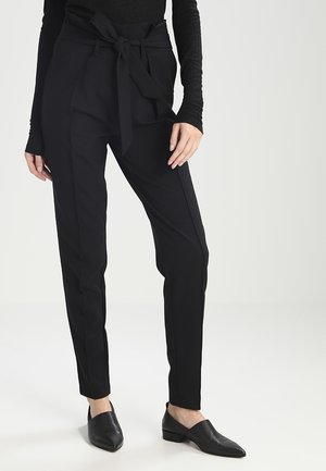 Frau trägt schwarze High-Waist-Paperbag-Hose mit gebundenem Gürtel und schwarze spitze flache Schuhe, steht vor einem schlichten Hintergrund.
