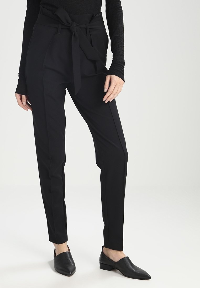 Femme portant un pantalon paperbag noir taille haute avec une ceinture nouée et des chaussures plates noires pointues, debout devant un fond uni.