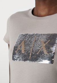 Camiseta de algodón beige con un parche rectangular de lentejuelas plateadas con letras doradas "A/X". Diseño de mangas cortas y cuello redondo.