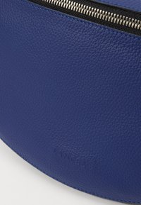 Zila ādas soma ar teksturētu virsmu, melnu rāvējslēdzēju un izsistām logotipu. Gludas malas papildina tās apaļo formu.