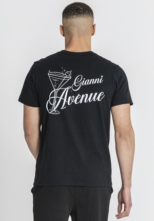 Mand med kort hår iført en sort t-shirt med hvid tekst "Gianni Avenue" og en grafik af et martiniglas på ryggen.