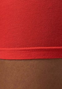 Tessuto di cotone rosso con una texture liscia, caratterizzato da un bordo orlato. Lo sfondo rivela un tono di pelle marrone chiaro.