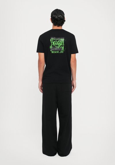 Black cotton T-shirt viewed from the back, featuring a neon green graphic design and text. Paired with black, wide-leg trousers.