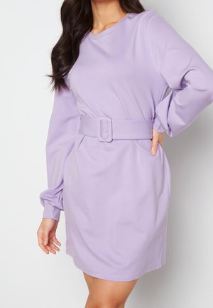 Femme portant une robe longue lilas à manches longues bouffantes, ceinturée, main sur la hanche, sur un fond uni clair.