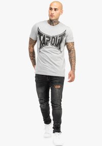 Pelēka kokvilnas T-kreklu ar lielu melnu "TAPOUT" logo, apvienojot ar nolietotām melnām džinsām un baltām sporta kurpēm.