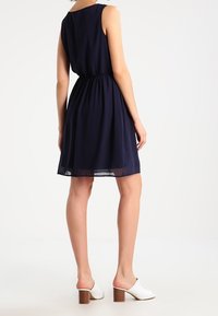 Marinblå ärmlös klänning med samlad midja, utsvängd kjol och genomskinlig textur. Matchad med vita klackskor med blockhæl.