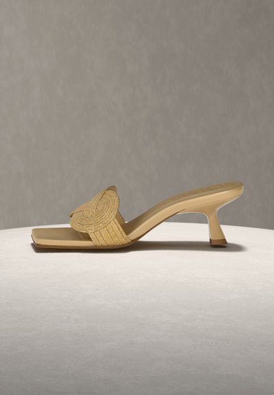 Bézs színű, négyzet alakú orrú, fonott pántos magassarkú papucs, világos, texturált felületen, semleges háttér előtt bemutatva.