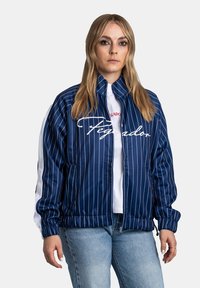 Granatowa marynarka w paski z zamkiem błyskawicznym, elastycznymi mankietami i pasem w talii. Posiada białe akcenty oraz haftowane logo "Pegador".