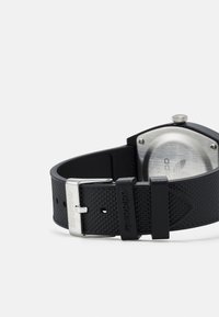 Montre connectée noire en caoutchouc avec un bracelet texturé, des éléments en métal argenté, et un cadran carré minimaliste affichant un dos métallique et une couronne.