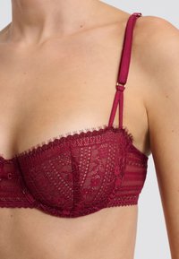 Reggiseno in pizzo bordeaux con tessuto testurizzato, delicati motivi floreali e spalline regolabili. Caratterizzato da bordi ondulati e chiusura a gancio.