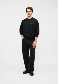 Μαύρο βαμβακερό φούτερ με στρογγυλή λαιμόκοψη και μανίκια με μανσέτα, συνδυασμένο με φαρδιά μαύρα τζιν και μαύρα αθλητικά παπούτσια. Λεπτομέρεια με λογότυπο στο στήθος.