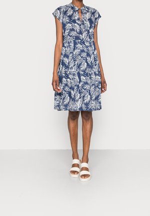 Kobieta w granatowej sukience do kolan z białym wzorem liści, krótkimi rękawami, dekoltem z wycięciem i białymi sandałami na platformie.