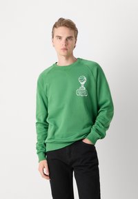 Grön sweatshirt i bomull med ribbade ärmslut, med en vit grafisk tryck på framsidan och rundad halsringning. I kombination med svarta byxor.