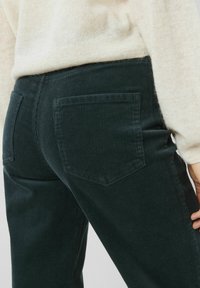 Pantalon en velours côtelé vert foncé avec des côtes verticales, comportant deux poches arrière et une coupe ajustée, associé à un pull de couleur claire.