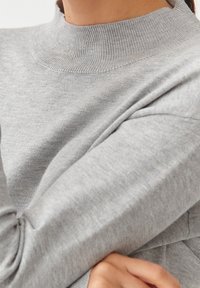 Maglione grigio chiaro con un collo alto a coste e un tessuto morbido e strutturato. Le maniche sono leggermente larghe e aderenti ai polsi.