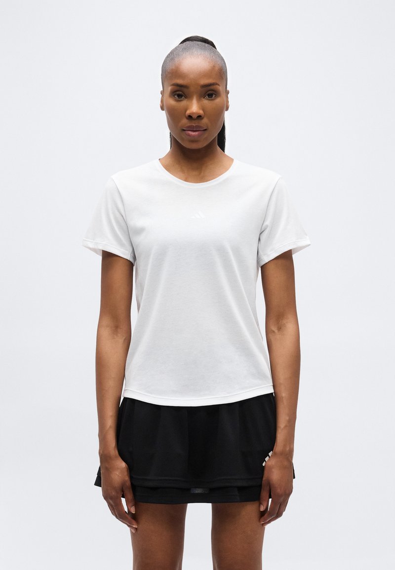 Γυναίκα με σκούρο δέρμα και μαλλιά δεμένα πίσω, που στέκεται φορώντας ένα απλό λευκό T-shirt και μια μαύρη φούστα μπροστά σε ένα απλό φωτεινό φόντο.