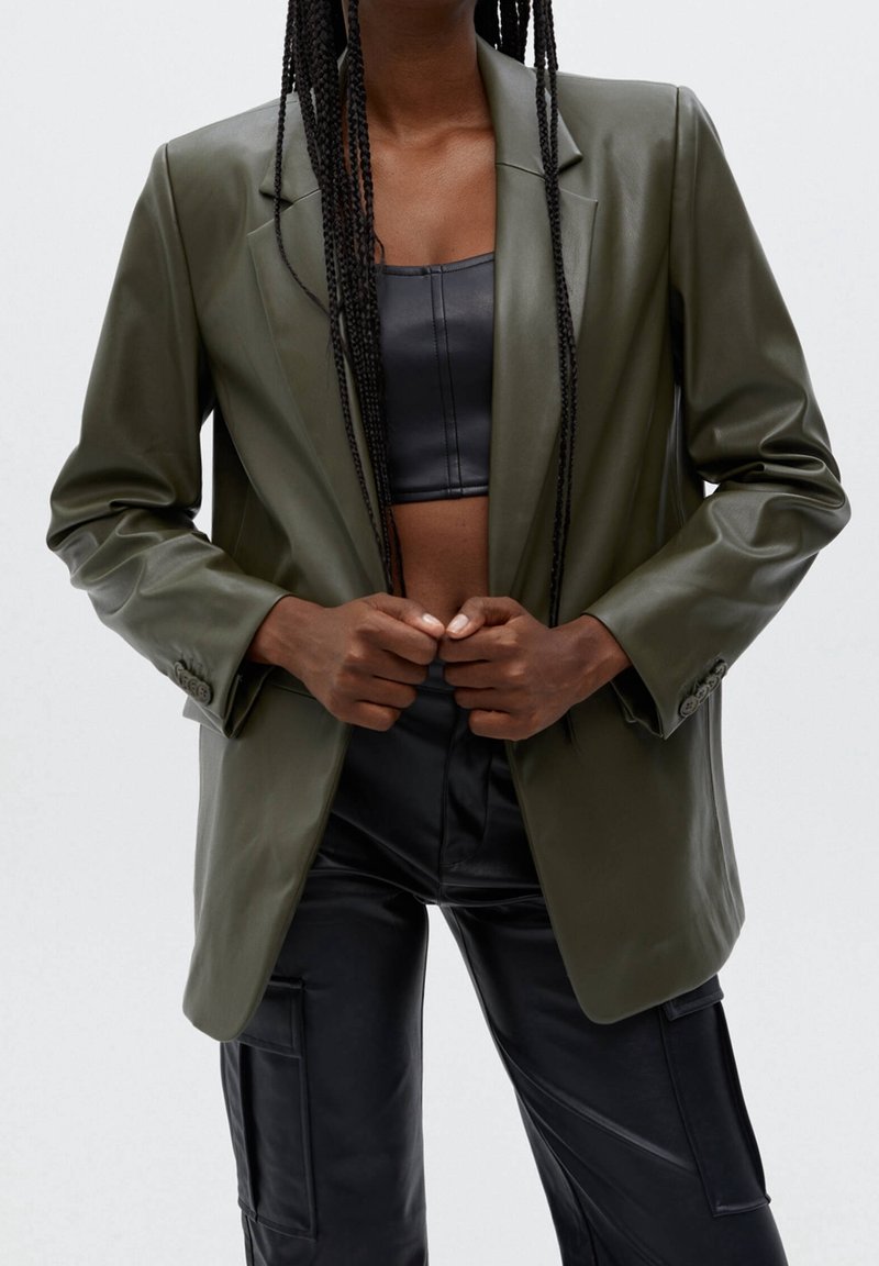 Blazer en cuir vert olive avec un col à revers cranté, une fermeture à bouton unique et une coupe décontractée, associé à un top court en cuir noir et un pantalon noir.
