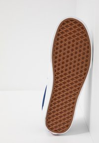 Sohle eines Sneakers mit braunem Waffelmuster-Gummi-Außensohle und weißem Zwischensohl, vertikal gegen weiße Flächen positioniert.