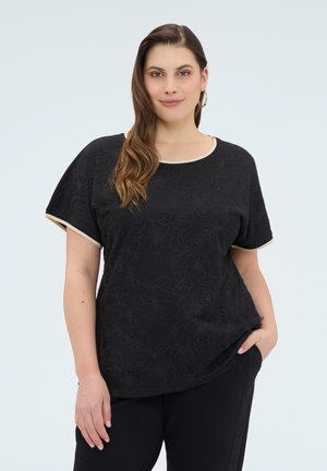 Frau mit langen braunen Haaren, die ein schwarzes Oberteil mit Blumenmuster und cremefarbenem Saum sowie passende schwarze Hosen trägt, steht mit einer Hand in der Tasche.