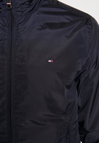 Marineblauwe jas van glanzende, gladde stof met een ritssluiting aan de voorkant. Bevat een geborduurd logo met rode, witte en blauwe accenten.